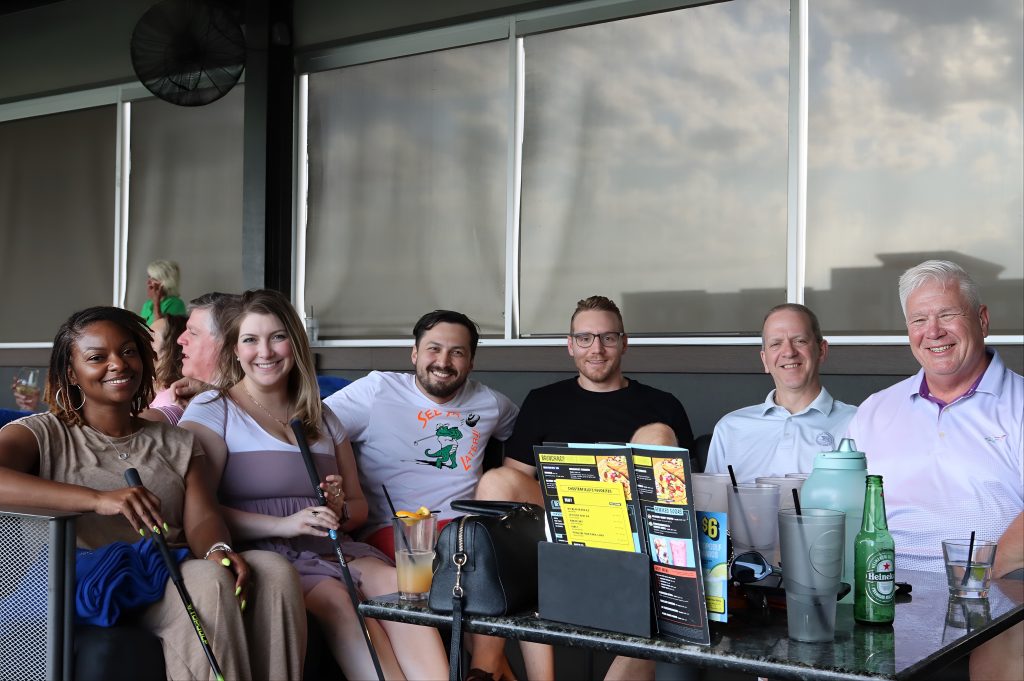 Diverse group of 4 men and 3 women sitting at a table at Top Golf