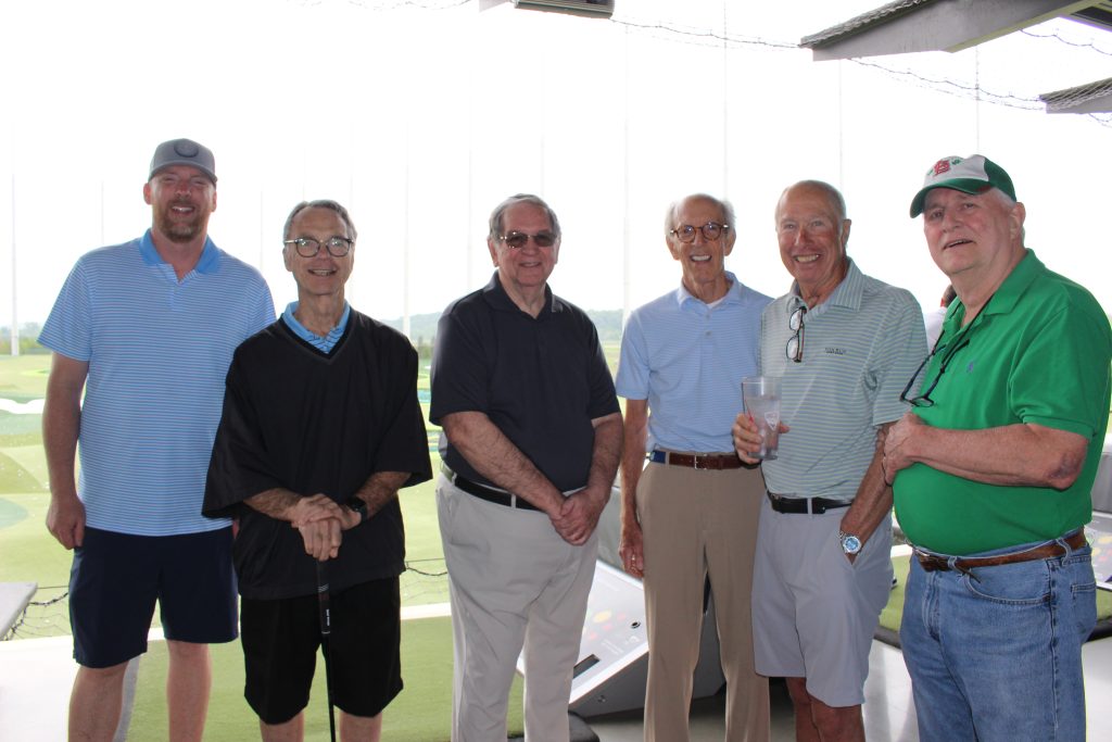 group of six older gentlemen in front of the Top Golf course smiling